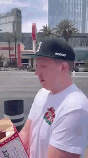 a man wearing a hat and a white t-shirt is holding a box of food .