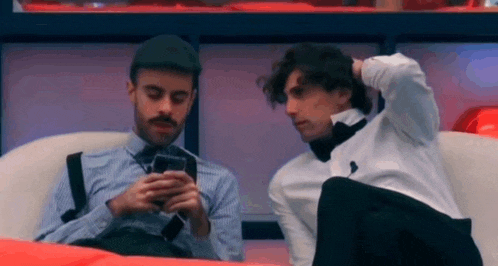 two men are sitting next to each other on a couch and one of them is looking at his phone .