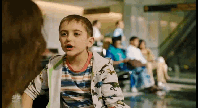 a young boy wearing a dinosaur shirt is talking to a woman in an airport waiting area .