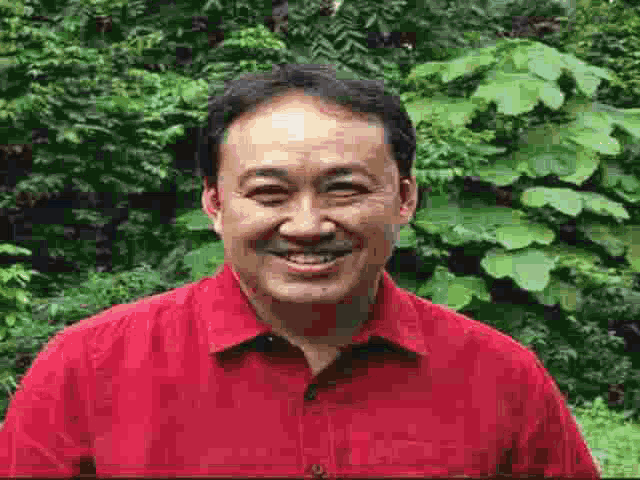 a man in a red shirt is smiling for the camera in front of trees .