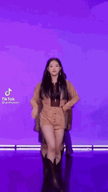 a woman in a brown jacket and shorts is dancing on a stage against a purple background .