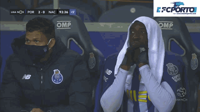 a man wearing a mask sits next to a man with a towel wrapped around his head in front of a omp seat