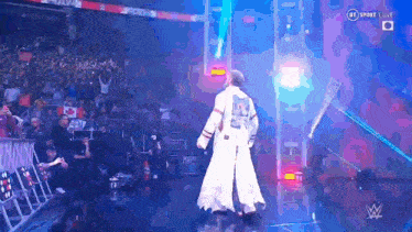 a man in a white robe with the letter r on his back walks into a wrestling ring