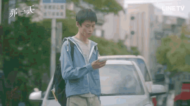 a man in a blue jacket is standing in front of a car looking at his cell phone .