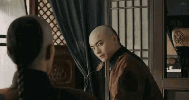 a man with a shaved head is standing in front of a window looking at another man .