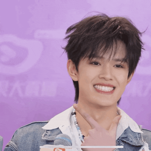 a young man wearing a necklace and a denim jacket smiles in front of a purple background