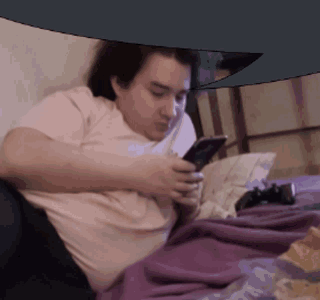 a man in a pink shirt is laying on a bed looking at his phone