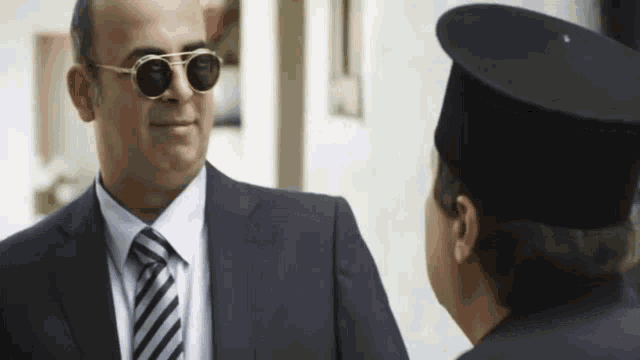 a man in a suit and tie wearing sunglasses talks to another man in a hat