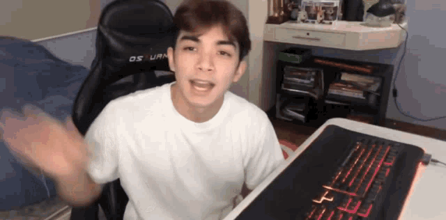 a young man in a white shirt is sitting at a desk with a keyboard and a black chair that says os uran