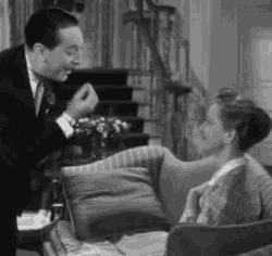 a man in a suit and tie is standing next to a woman sitting on a couch .