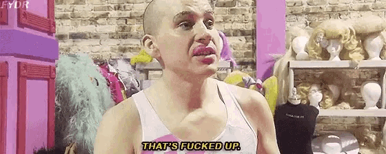 a bald man with pink lipstick on his lips is standing in front of a brick wall and wigs .