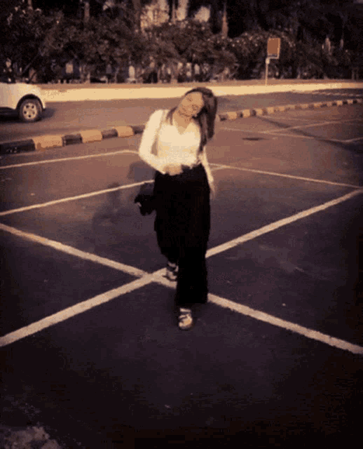 a woman in a white shirt and black pants is standing in a parking lot with a white car in the background