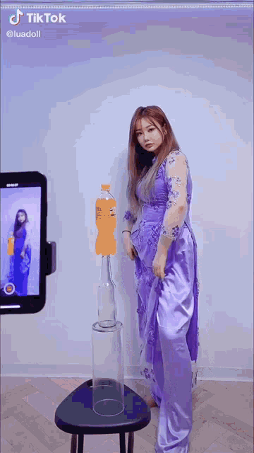 a woman in a purple dress is standing next to a bottle of orange soda and a glass