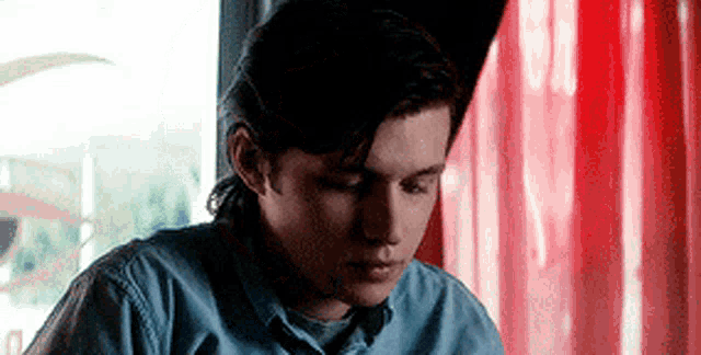 a young man in a blue shirt is sitting at a table with a red curtain in the background .