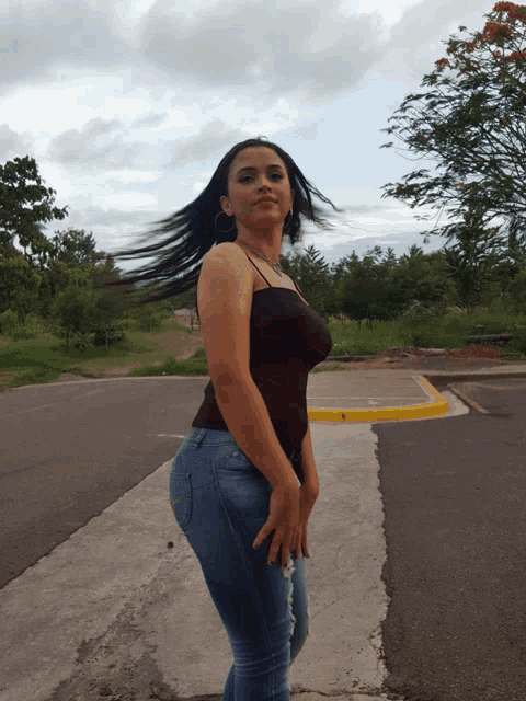 a woman in a black tank top and blue jeans stands on a street