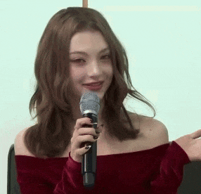 a woman is holding a microphone and smiling while wearing a red off the shoulder top .