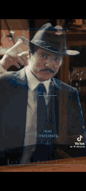 a man in a suit and hat is wearing glasses and a mustache and has the word conference on the screen behind him