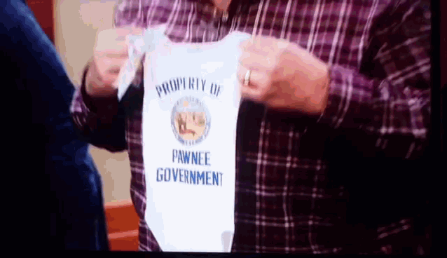 a man in a plaid shirt is holding a white baby onesie that says " property of pawnee government "