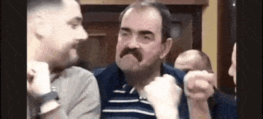 a man with a mustache is sitting in a room with other men and making a funny face .