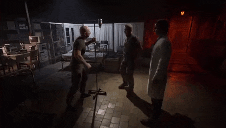a man in a white coat is standing in a dark room talking to two other men .