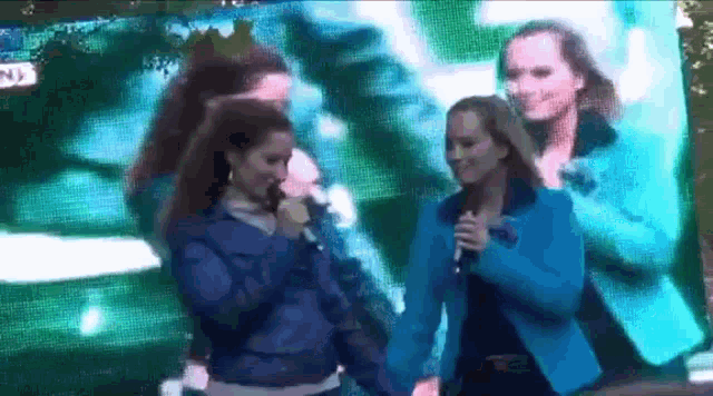 two women in blue jackets are singing into microphones on a stage .