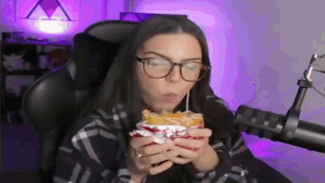 a woman wearing glasses is sitting in front of a microphone eating a hamburger .