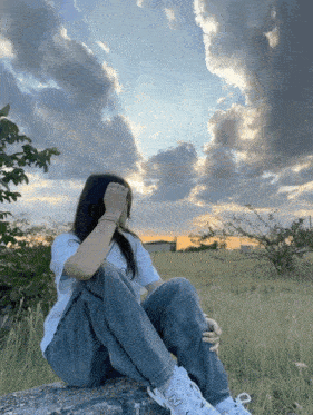 a woman sitting on a rock in a field with a cloudy sky in the background