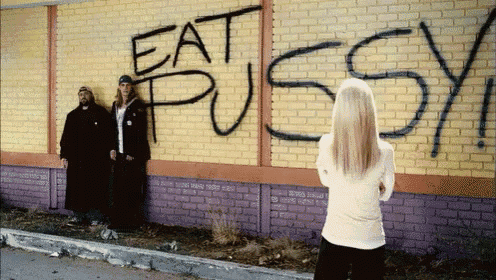 a woman stands in front of a brick wall that has the word eat pussy spray painted on it