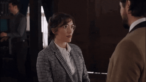a woman wearing glasses and a plaid jacket looks surprised while talking to a man in a suit