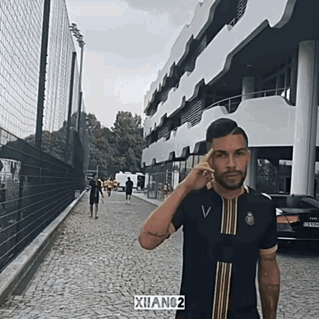 a man wearing a black shirt with gold stripes is standing in front of a building that says xiano2