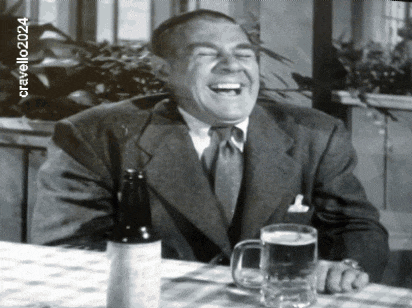 a man in a suit and tie is laughing while sitting at a table with a beer mug and a bottle of beer