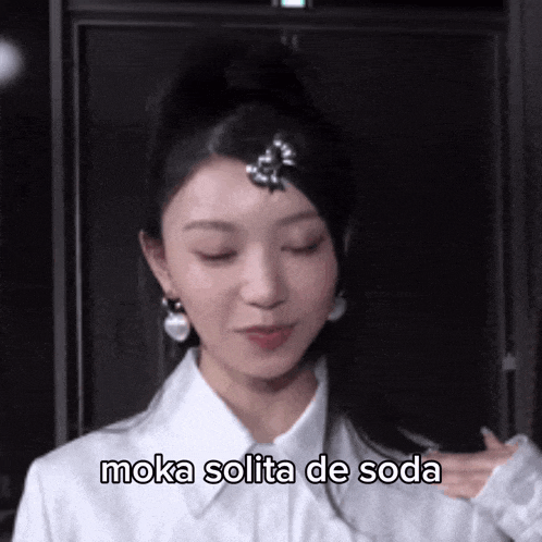 a woman wearing a white shirt and earrings is making a funny face with the words moka solita de soda above her head .