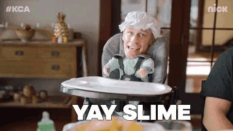 a man is sitting in a high chair with a baby doll on his head and the words `` yay slime '' .