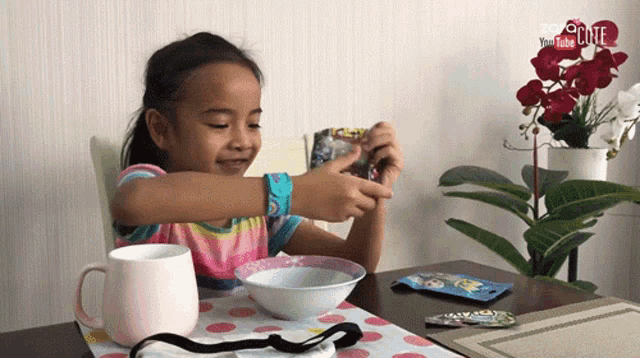 a little girl sits at a table with a cup and a bowl and a youtube logo behind her