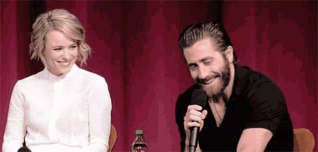 a man with a beard is talking into a microphone while sitting next to a woman