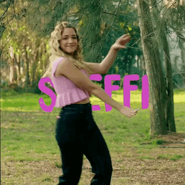 a woman in a pink top and black jeans is dancing in front of a sign that says stiff