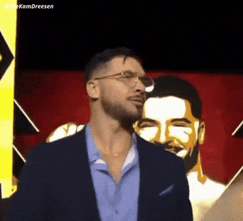 a man wearing glasses and a suit is standing in front of a painting of a man with his eyes closed