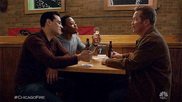 three men are sitting at a table with bottles of beer and a sign that says happy hour on it