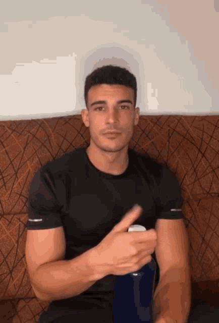 a man in a black shirt is sitting on a couch holding a blue bottle and giving a thumbs up