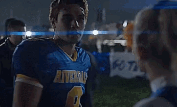 a man in a riverdale football jersey stands in front of cheerleaders .