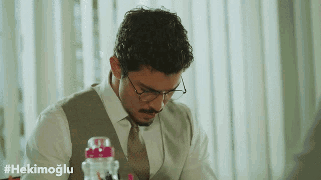 a man wearing glasses sits at a desk with the hashtag #hekimoglu on the bottom right