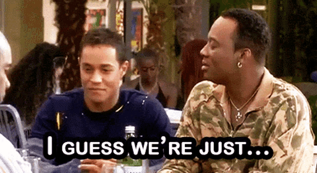 two men are sitting at a table and one of them is saying " i guess we 're just " .