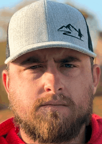 a man with a beard is wearing a hat with a mountain and a bird on it