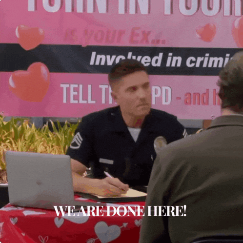 a man sitting at a table with a laptop and a sign that says " we are done here " behind him