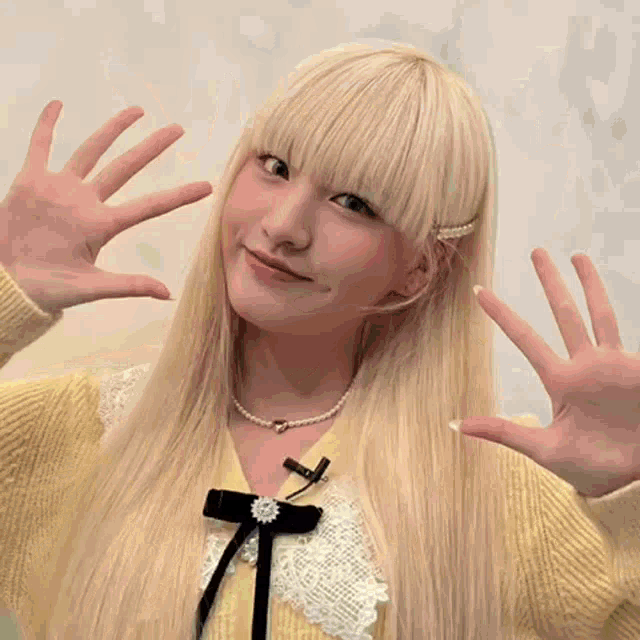 a blonde girl wearing a yellow sweater and a pearl necklace is making a face with her hands .