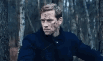a man in a blue jacket is standing in the woods with blood on his face .