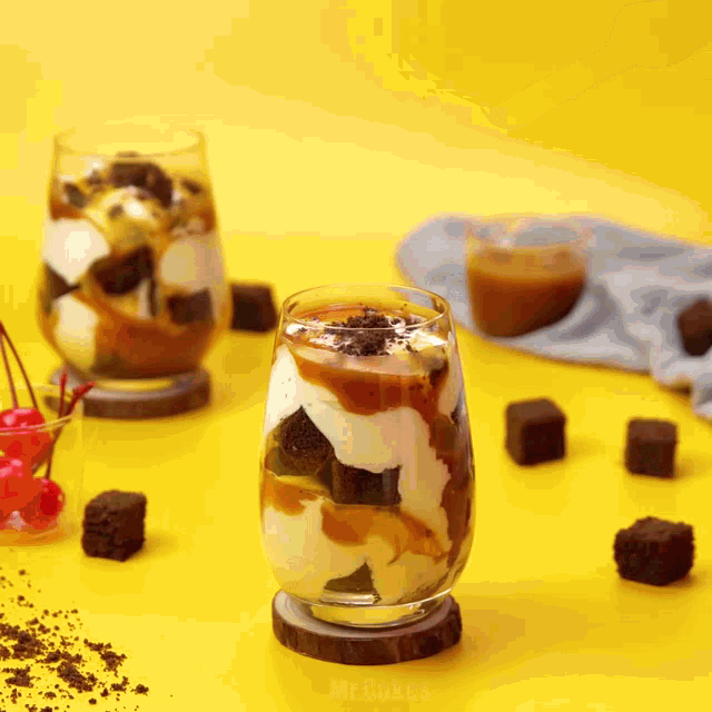 two glasses filled with ice cream and brownies are on a yellow surface