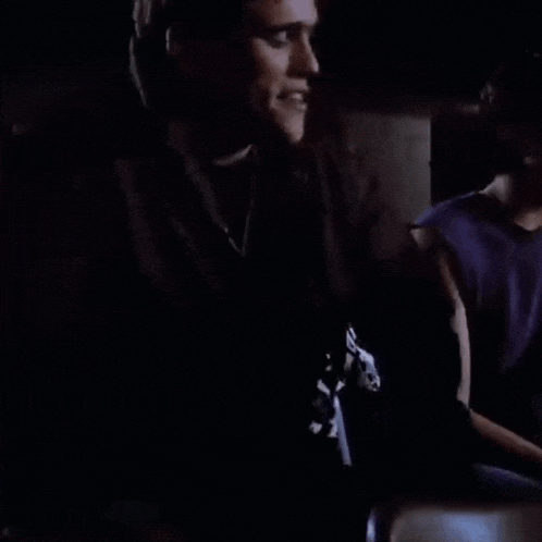 a man in a black jacket is sitting in a dark room talking to another man .