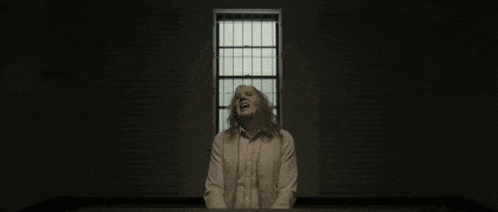 a man in a white shirt is sitting in front of a window in a dark room