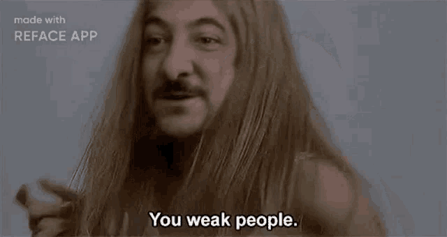 a man with long hair and a mustache is standing in front of a wall and saying `` you weak people . ''